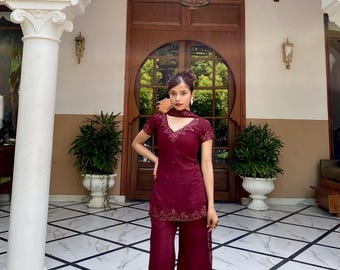 May include: A deep burgundy two-piece outfit consisting of a short-sleeved tunic with a V-neckline and wide-leg trousers. The tunic and trousers feature intricate gold embroidery along the hemlines and neckline, paired with a matching scarf.