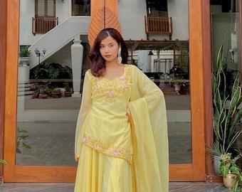May include: A bright yellow three-piece ensemble consisting of a long-sleeved tunic with pink floral embroidery at the neckline and hem, a matching floor-length pleated skirt, and a sheer yellow dupatta draped over the shoulder.