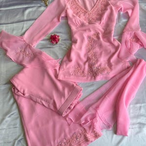 May include: A light pink long-sleeved tunic and matching trousers set featuring intricate tonal embroidery and beadwork along the V-neckline, bodice, and sleeve cuffs. The sleeves have flared, layered ruffles, and the trousers feature a wide-leg design.