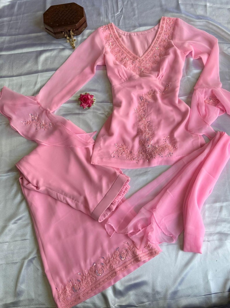 May include: A light pink long-sleeved tunic and matching trousers set featuring intricate tonal embroidery and beadwork along the V-neckline, bodice, and sleeve cuffs. The sleeves have flared, layered ruffles, and the trousers feature a wide-leg design.