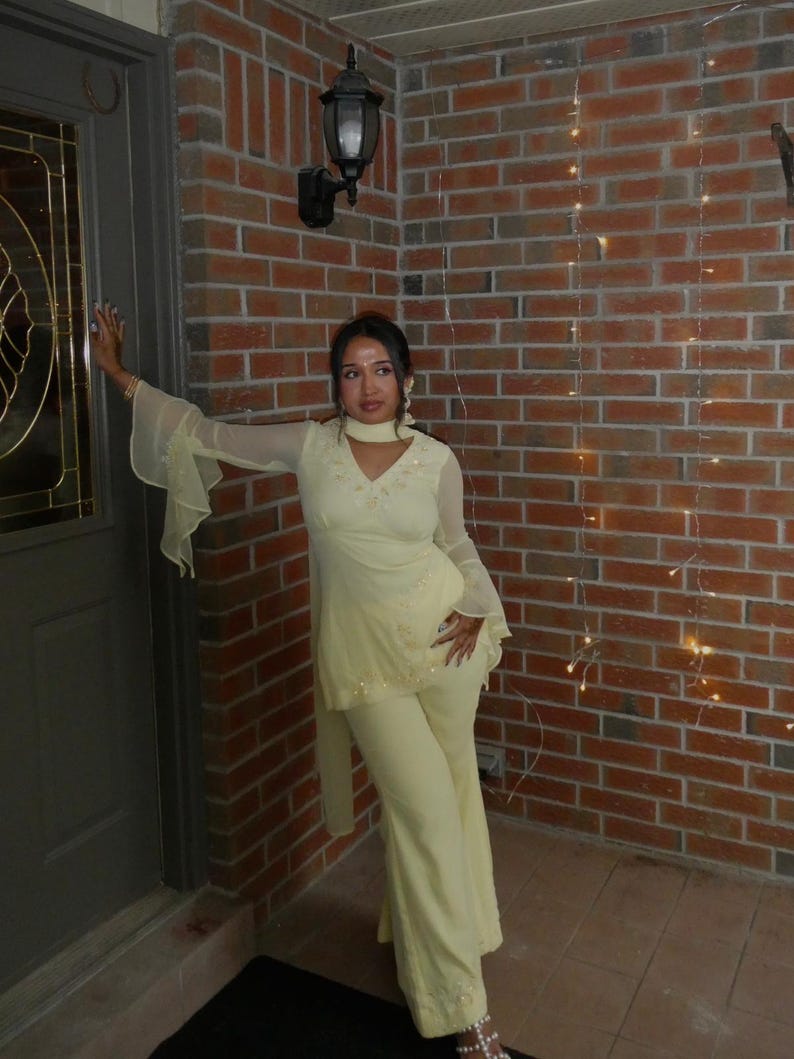 May include: A pale yellow two-piece outfit consisting of a long-sleeved tunic with flared cuffs, a V-neckline, and matching wide-leg trousers. The tunic features delicate floral embroidery along the neckline and hem, with a coordinating scarf neck wrap.