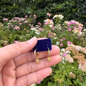 Può includere: Un paio di orecchini a cerchio dorati con piccoli ciondoli a forma di campana incastonati con minuscole pietre trasparenti. Gli orecchini sono fissati su un piccolo pezzo rettangolare di tessuto di velluto blu scuro.