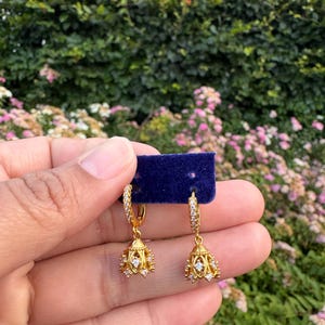 Puede incluir: Un par de pendientes de aro dorados con un dije en forma de campana. Cada aro está incrustado con pequeñas piedras transparentes, y el colgante en forma de campana tiene un diseño de filigrana con piedras adicionales, sobre una tarjeta azul.