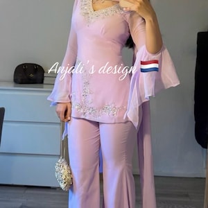 May include: A light purple two-piece outfit consisting of a long-sleeved tunic with flared sleeves and wide-leg trousers. The tunic features silver embroidery around the V-neckline and hem. The text Anjali's design is visible on the garment.
