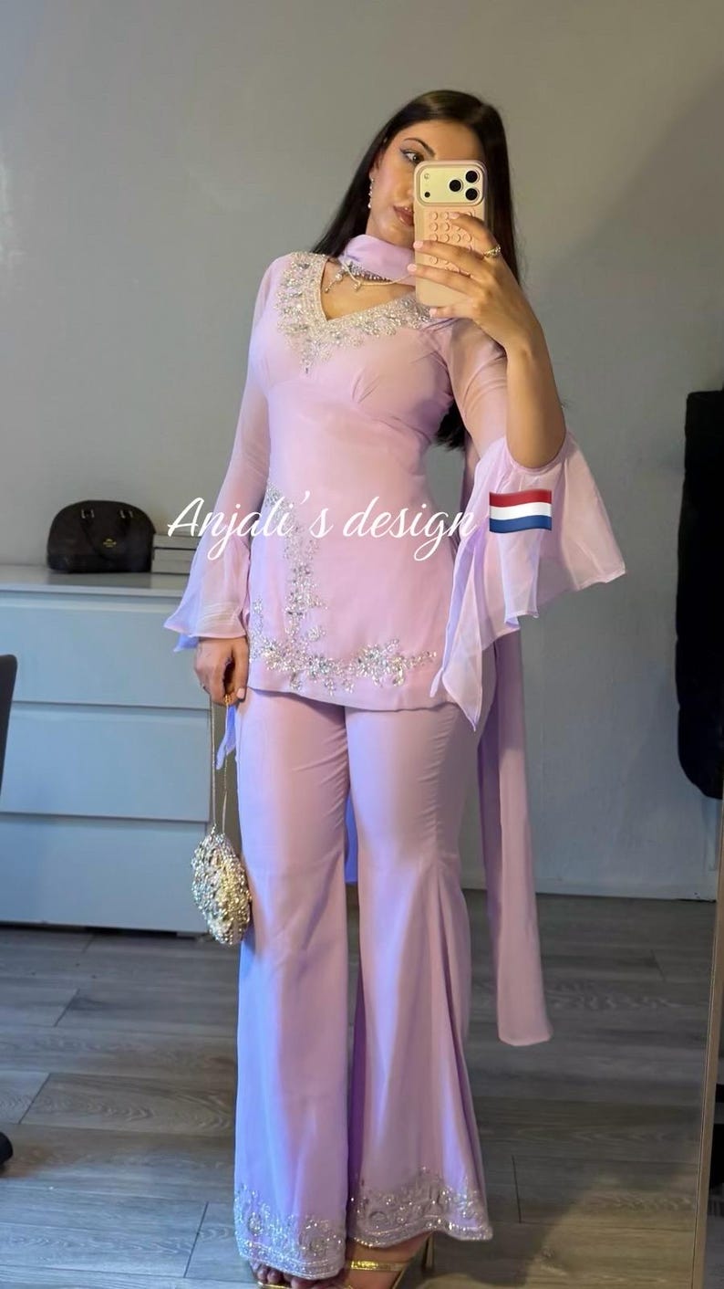 May include: A light purple two-piece outfit consisting of a long-sleeved tunic with flared sleeves and wide-leg trousers. The tunic features silver embroidery around the V-neckline and hem. The text Anjali's design is visible on the garment.