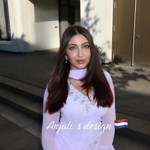 May include: A light lavender long-sleeved tunic featuring a V-neckline with silver sequin embellishments, flared cuffs, and a matching scarf. The text Anjali's design is printed in white script next to a small Dutch flag icon on the garment.