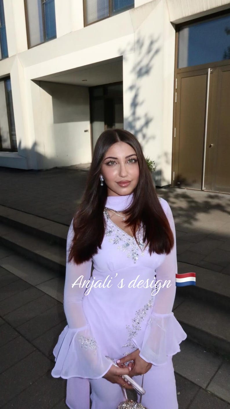 May include: A light lavender long-sleeved tunic featuring a V-neckline with silver sequin embellishments, flared cuffs, and a matching scarf. The text Anjali's design is printed in white script next to a small Dutch flag icon on the garment.
