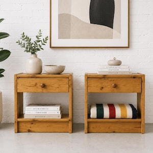 May include: Two wooden nightstands with drawers and lower shelves. Each nightstand has a round wooden knob on the drawer. One nightstand has a folded blanket with red, green, and yellow stripes. A potted plant and artwork are in the background.