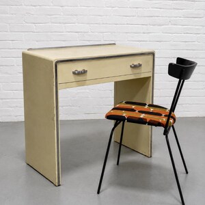 May include: A cream-colored desk with a single drawer and metallic trim, paired with a black metal chair featuring a seat upholstered in a geometric pattern of orange, brown, black, and white shapes. The desk has a smooth, flat top surface.