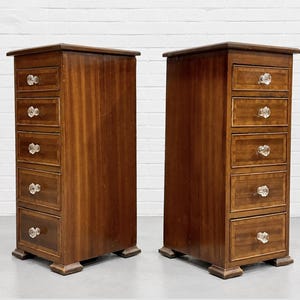 May include: A pair of tall, narrow wooden chests of drawers, each featuring five stacked drawers with clear glass knobs. The wood has a medium brown finish with lighter wood inlay borders around each drawer front and a sturdy base with feet.