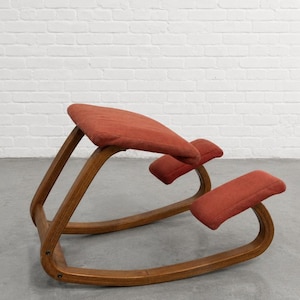May include: An ergonomic kneeling chair featuring a curved wooden frame with a natural brown finish. The chair includes a padded seat and a separate padded knee rest, both upholstered in a textured, burnt orange fabric for support.