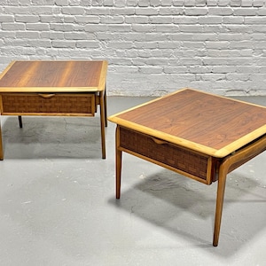 May include: Two square mid-century modern side tables. Each table has a dark wood top with a light wood border, a woven drawer, and angled wooden legs. The tables are set against a white brick wall.