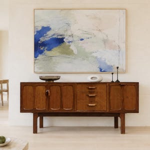 May include: A rectangular abstract painting in a light wood frame featuring blue, olive green, and cream brushstrokes with thin black lines. The artwork hangs above a dark brown wooden sideboard with two cabinet doors and three central drawers.