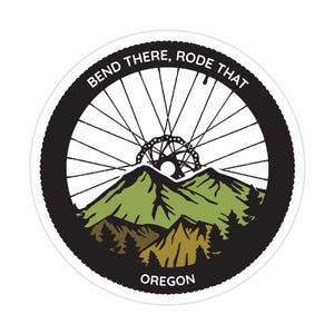 May include: Black and white sticker with a mountain landscape and a bicycle wheel in the background. The text "Bend There, Rode That Oregon" is printed on the sticker.