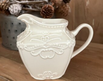 May include: Off-white ceramic creamer with an ornate, embossed design. The creamer features a spout, a handle, and decorative detailing, including floral and beaded patterns. The creamer is set against a blurred background.