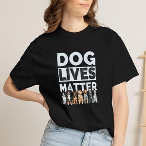 以下が含まれることがあります： 胸の中央にDOG LIVES MATTERという白いテキストがブロック体で重ねてプリントされ、その下に7種類の異なる犬種が並んでいるカラフルなグラフィックイラストが描かれた、黒い半袖のTシャツ。
