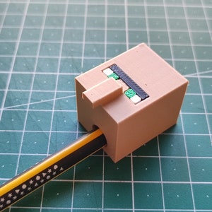 Peut inclure: Un porte-crayon brun en forme de tête de personnage Minecraft. Le porte-crayon a une bouche noire et des yeux verts. Un crayon jaune est inséré dans le porte-crayon.