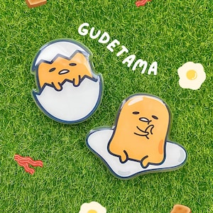 May include: Two Gudetama character pins, one in an eggshell and the other on a fried egg, with the word "Gudetama" above them. The image also includes bacon, toast, and fried egg stickers on a green background.