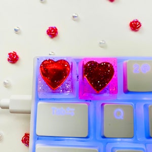 Pu&ograve; includere: Due tasti a forma di cuore con glitter rosso incorporati in resina trasparente. I tasti sono su una tastiera bianca con retroilluminazione blu. La tastiera ha tasti etichettati "Tab", "Bloc Maiusc", "Q" e "A".