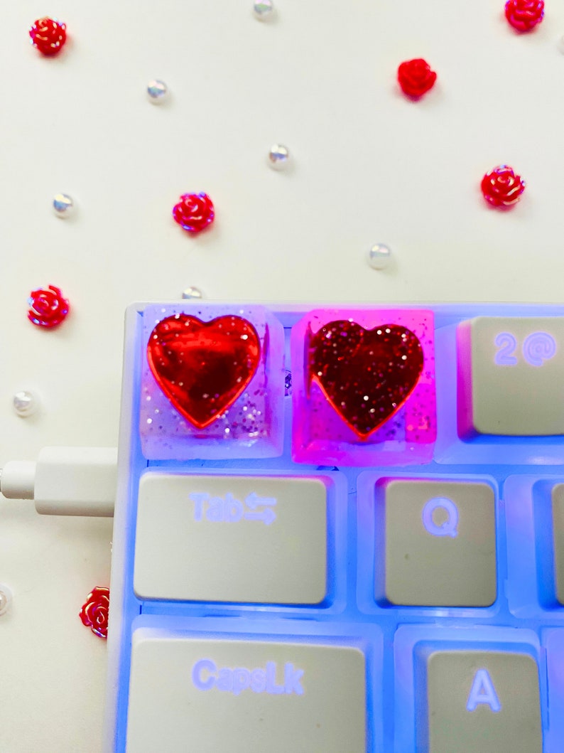 Pu&ograve; includere: Due tasti a forma di cuore con glitter rosso incorporati in resina trasparente. I tasti sono su una tastiera bianca con retroilluminazione blu. La tastiera ha tasti etichettati "Tab", "Bloc Maiusc", "Q" e "A".