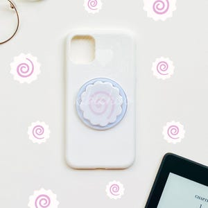 May include: White phone case with a light purple and white pop socket featuring a spiral design. The image includes decorative spiral stickers and a tablet with text. Gold-rimmed glasses are also visible.