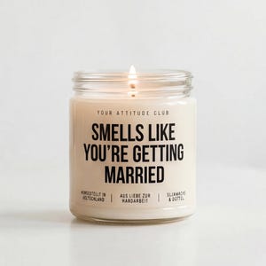 Könnte beinhalten: Eine brennende, cremefarbene Kerze in einem Glas. Auf der Kerze steht in schwarzer Schrift "SMELLS LIKE YOU'RE GETTING MARRIED". Das Glas trägt auch den Text "YOUR ATTITUDE CLUB" und weiteren Text auf Deutsch.