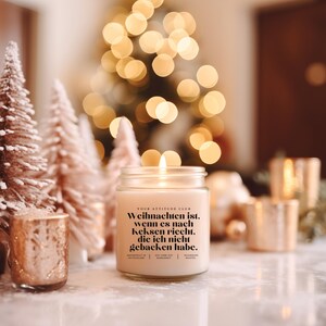 Può includere: Una candela accesa in un barattolo di vetro con il testo "YOUR ATTITUDE CLUB" e una citazione tedesca sul Natale. La candela è circondata da piccoli alberi di Natale e altri oggetti decorativi, con un albero di Natale sfocato sullo sfondo.