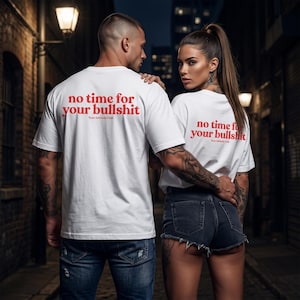 Peut inclure: Deux personnes portant des t-shirts blancs avec le texte rouge "no time for your bullshit" et "Your Attitude Club" imprimé au dos. L'arrière-plan est une ruelle sombre avec des lampadaires.