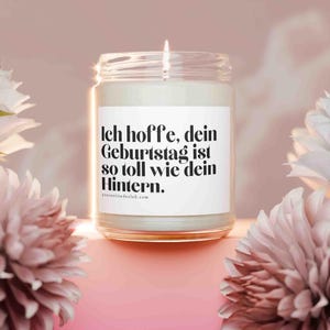 Könnte beinhalten: Eine brennende Kerze in einem durchsichtigen Glas mit einem weißen Etikett. Das Etikett trägt den schwarzen Text: "Ich hoffe, dein Geburtstag ist so toll wie dein Hintern." Der Hintergrund ist rosa mit weißen und rosa Blumen.