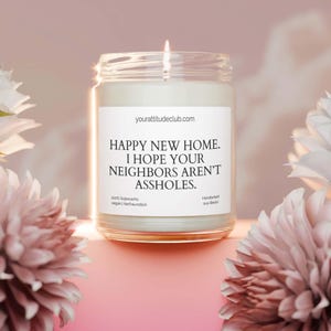Może przedstawiać: Zapalone świeca w przezroczystym szklanym słoiku z białą etykietą. Na etykiecie widnieje napis "HAPPY NEW HOME. I HOPE YOUR NEIGHBORS AREN'T ASSHOLES." Strona internetowa yourattitudeclub.com jest wydrukowana nad tekstem. Świeca otoczona jest różowymi i białymi kwiatami.