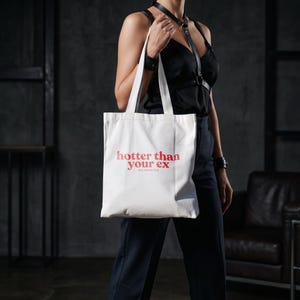 Könnte beinhalten: Eine weiße Tragetasche mit dem Text "hotter than your ex" in Rot. Die Tasche wird von einer Person gehalten. Der Hintergrund ist eine dunkelgraue Wand.