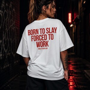 Könnte beinhalten: Ein weißes kurzärmeliges T-Shirt mit rotem Text auf der Rückseite, der BORN TO SLAY FORCED TO WORK lautet, und kleinerem Text darunter, der YOUR ATTITUDE CLUB lautet. Das T-Shirt hat eine lockere Passform und wird mit einer schwarzen Cargohose kombiniert.