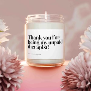 Könnte beinhalten: Eine brennende Kerze in einem transparenten Glas mit einem weißen Etikett. Auf dem Etikett steht "Thank you for being my unpaid therapist!" in schwarzer Schrift. Der Hintergrund ist rosa mit weißen und rosa Blumen.