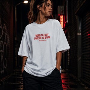 Könnte beinhalten: Ein weißes kurzärmeliges T-Shirt mit dem roten Schriftzug BORN TO SLAY FORCED TO WORK und dem kleineren Text YOUR ATTITUDE CLUB auf der Brust. Das Kleidungsstück hat einen Rundhalsausschnitt und eine lockere, kastige Passform mit weiten Ärmeln.