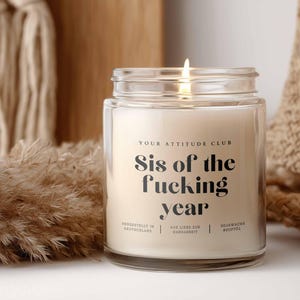 Könnte beinhalten: Eine brennende, klare Glas-Kerze mit dem Text "Sis of the fucking year" in Schwarz. Die Kerze ist von neutralfarbener Dekoration umgeben, darunter getrocknete Gräser und gewebte Texturen.