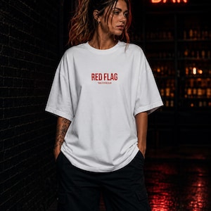 Könnte beinhalten: Ein weißes kurzärmeliges T-Shirt mit dem Text RED FLAG in fetten roten Großbuchstaben auf der Brust, wobei der kleinere Text YOUR ATTITUDE CLUB direkt darunter in einer passenden roten Schriftfarbe gedruckt ist.