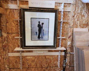 May include: A framed photograph of a couple dancing, with a dark frame and a light-colored mat. The photograph is mounted on a wooden shelf, with the name "SCOTT JACOBS" above it. The background is a wood-paneled wall.