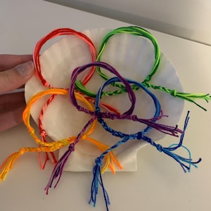 May include: A set of six colourful friendship bracelets with tassels. The bracelets are made of braided string in shades of red, orange, yellow, green, blue, and purple.