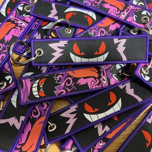 May include: A pile of purple and black keychains with a graphic of a ghost with red eyes and a white smile. The keychains are made of fabric and have a purple border.