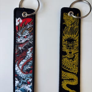 May include: Two black rectangular keychains with silver key rings. One keychain features a detailed dragon design in red, white, and blue against a red sky. The other keychain has a gold dragon design on a black background.