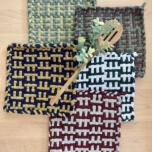 May include: Five woven pot holders in different colors and patterns. The pot holders are square and have a fringe border. A wooden spoon is laying on top of the pot holders.
