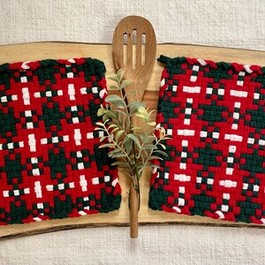 May include: Two square, woven pot holders with a red, green, and white plaid pattern. A wooden spoon and sprig of greenery are between the pot holders, all on a wooden cutting board.