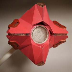 May include: A red, geometrically shaped, 3D printed model of a Ghost from the video game Destiny. The Ghost has a clear, circular window with a symbol inside.