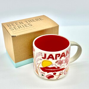 May include: Cream-colored ceramic mug with a red interior, featuring the word "JAPAN" and illustrations of Mount Fuji, cherry blossoms, and other Japanese icons. The mug is part of the "Been There Series" and comes with a cardboard box.