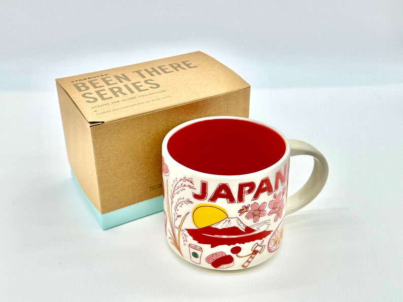 May include: Cream-colored ceramic mug with a red interior, featuring the word "JAPAN" and illustrations of Mount Fuji, cherry blossoms, and other Japanese icons. The mug is part of the "Been There Series" and comes with a cardboard box.