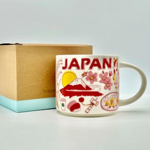 May include: A white ceramic mug with the word "JAPAN" in red lettering, featuring illustrations of Mount Fuji, cherry blossoms, and other Japanese icons. The mug is accompanied by a brown cardboard box with a light blue base.