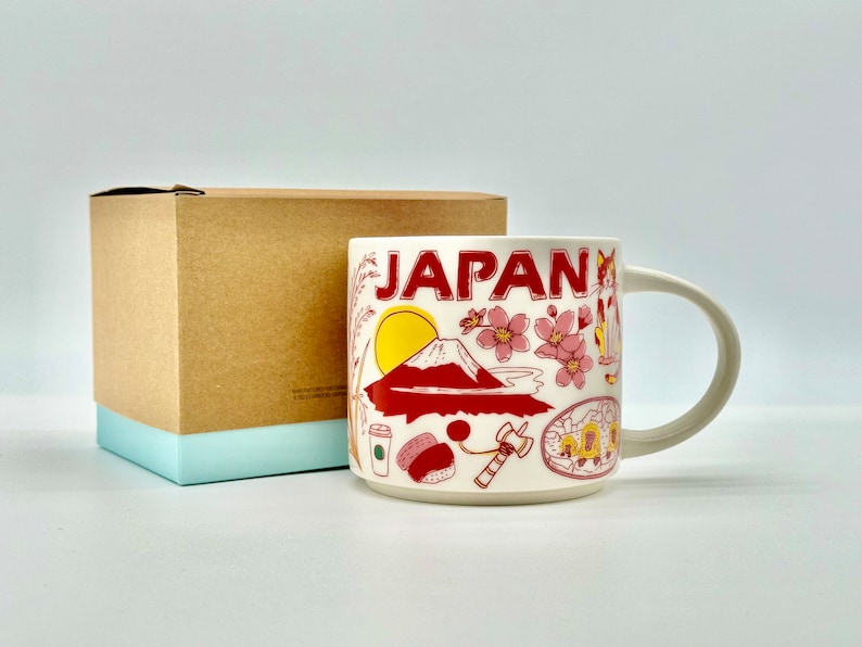 May include: A white ceramic mug with the word "JAPAN" in red lettering, featuring illustrations of Mount Fuji, cherry blossoms, and other Japanese icons. The mug is accompanied by a brown cardboard box with a light blue base.