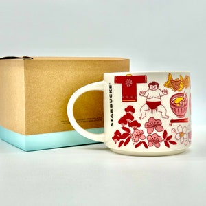 May include: White ceramic mug with a handle, featuring Japanese-themed illustrations in red and pink. The mug includes images of a sumo wrestler, cherry blossoms, and traditional Japanese food. The mug is next to a brown box with a light blue base.