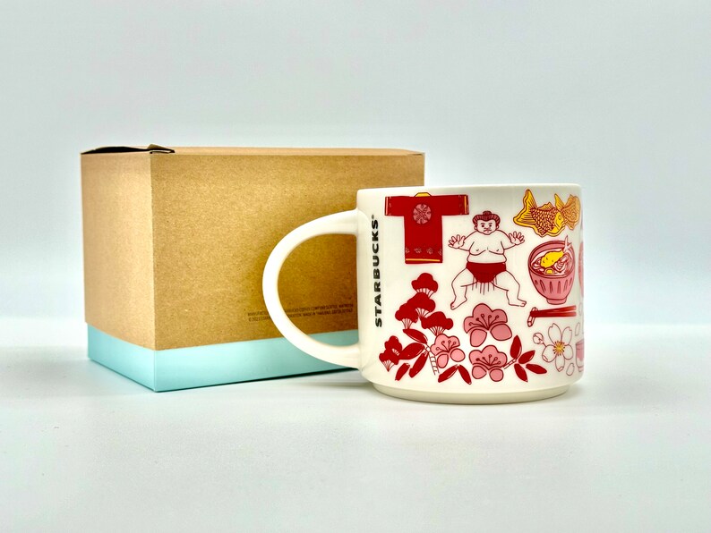 May include: White ceramic mug with a handle, featuring Japanese-themed illustrations in red and pink. The mug includes images of a sumo wrestler, cherry blossoms, and traditional Japanese food. The mug is next to a brown box with a light blue base.