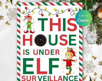 May include: A festive Christmas sign with the text "THIS HOUSE IS UNDER ELF SURVEILLANCE." The sign features a camera graphic, elf illustrations, and a border with red and green stripes. Decorative lights and Christmas ornaments surround the sign.